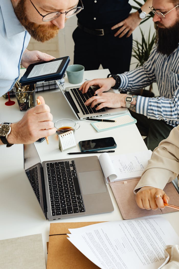 A Bearded Man Sitting at Desk Using a Laptop in Front of a Man Holding a Tablet